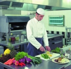 A fly killer unit on the wall behind a chef cutting vegetables in a commercial kitchen.